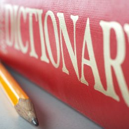 A red dictionary sitting on a table top with a pencil next to it.