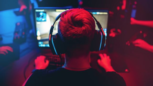Professional gamer shot from behind playing online games on computer with headphones on, on a blurred red and blue background.