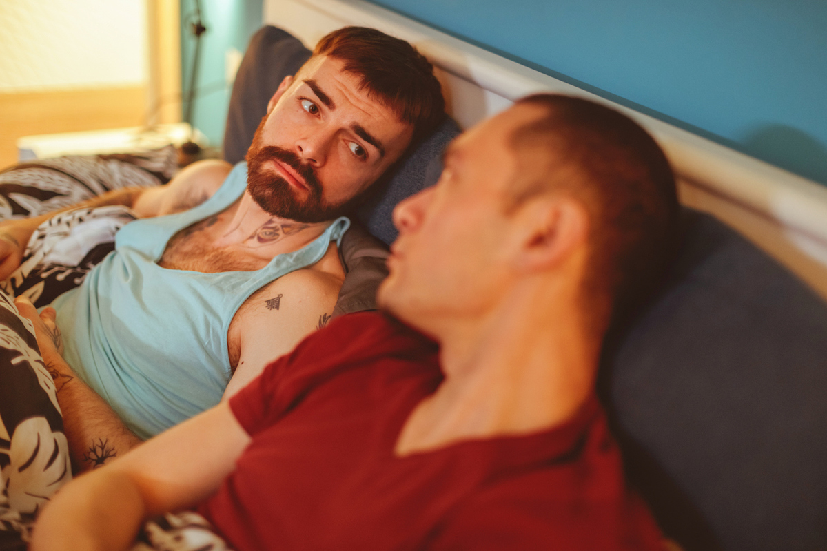 Sad man looking at his boyfriend lying in bed next to him