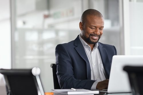 Положителен зрял бизнесмен, работещ на лаптоп в модерен офис.  Успешен африкански бизнесмен, работещ на компютър, докато седи на бюрото.  Усмихнат мъж на средна възраст, работещ в компания.