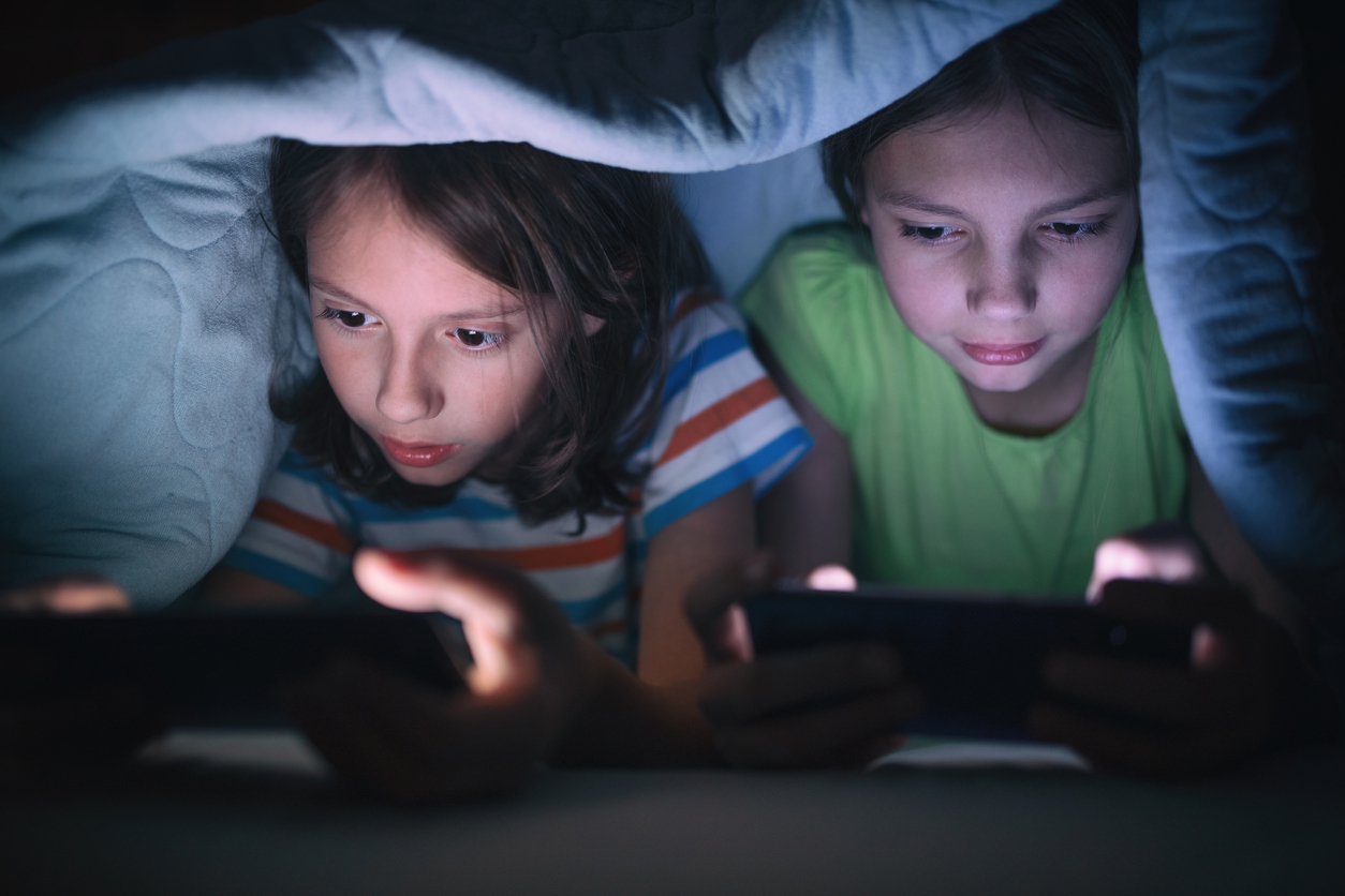 Two kids hiding under covers while playing video games on their phones.