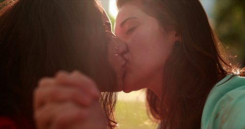 Women kissing outside while holding hands in the sun