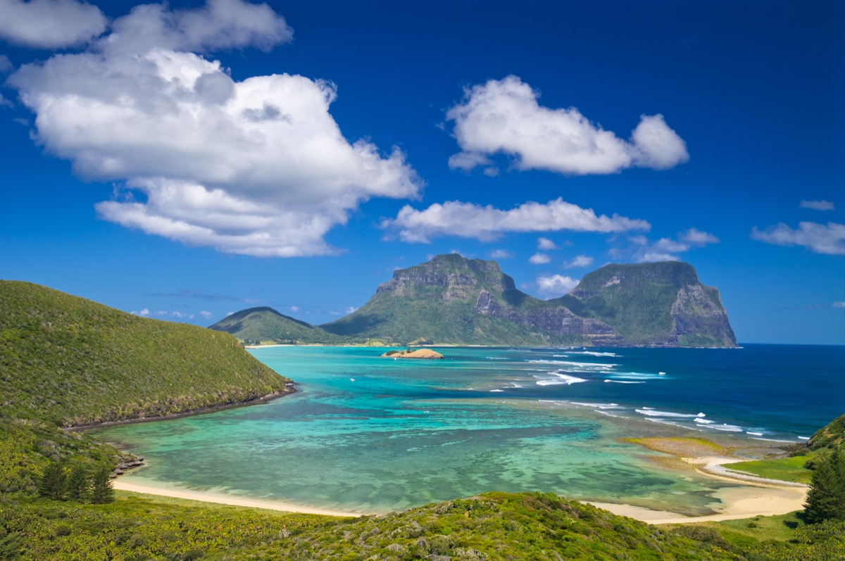View south over the stunning turquoise lagoon to the peaks of Mts Lidgbird and Gower. Taken from Mt Eliza on Lord Howe Island, Australia. Fluffy clouds stand out from the deep blue sky overhead. This tiny island is a delightful tourist destination.