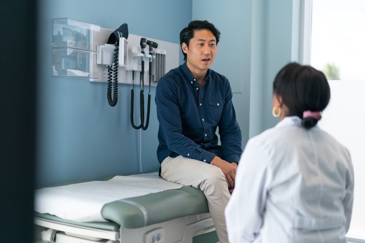 A man is at a routine medical appointment. The patient is sitting on an examination table facing his doctor. The kind doctor is listening as he speaks.