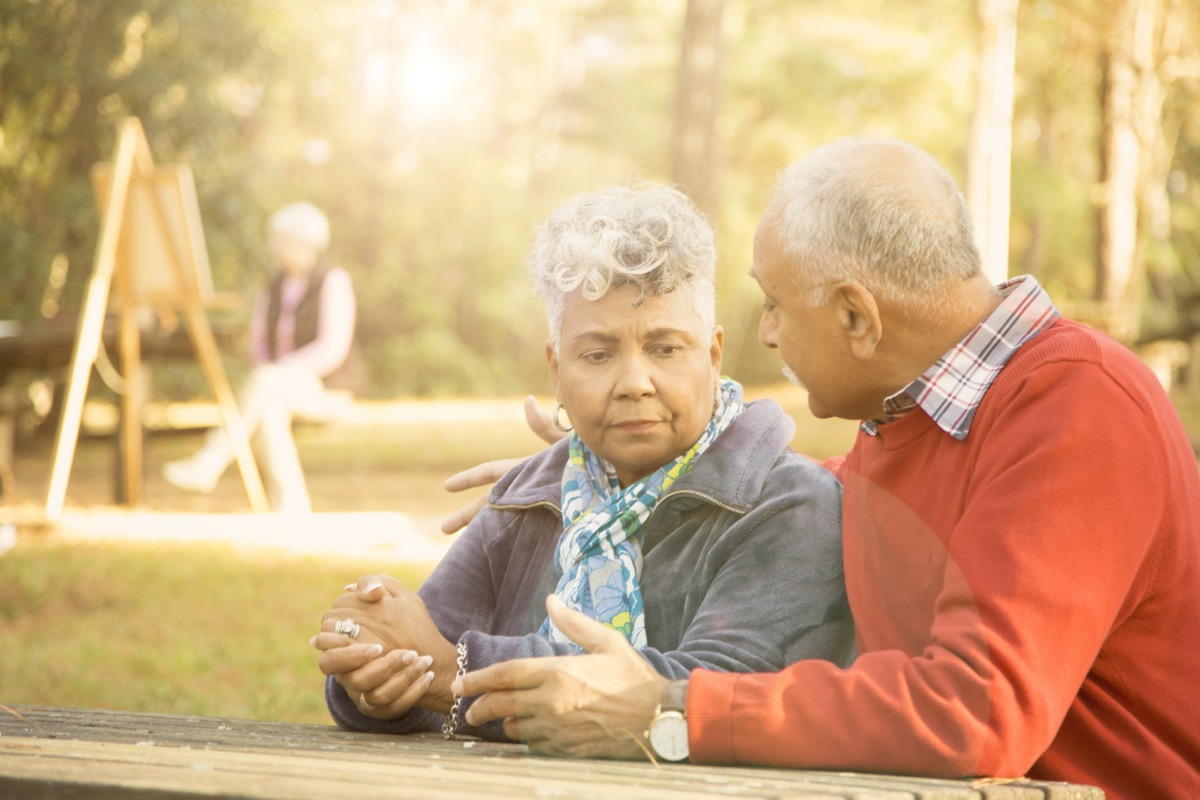 Mixed race senior adult couple talk at outdoor park in spring or autumn season. They sit on a park bench and discuss their relationship difficulties. African and middle eastern descent couple. Woman painting using easel in background.