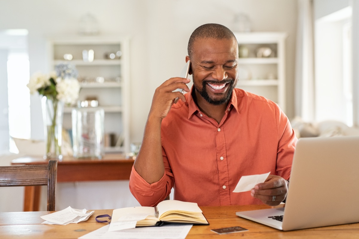 man sitting at table talking on phone and smiling, looking at paperwork and laptop