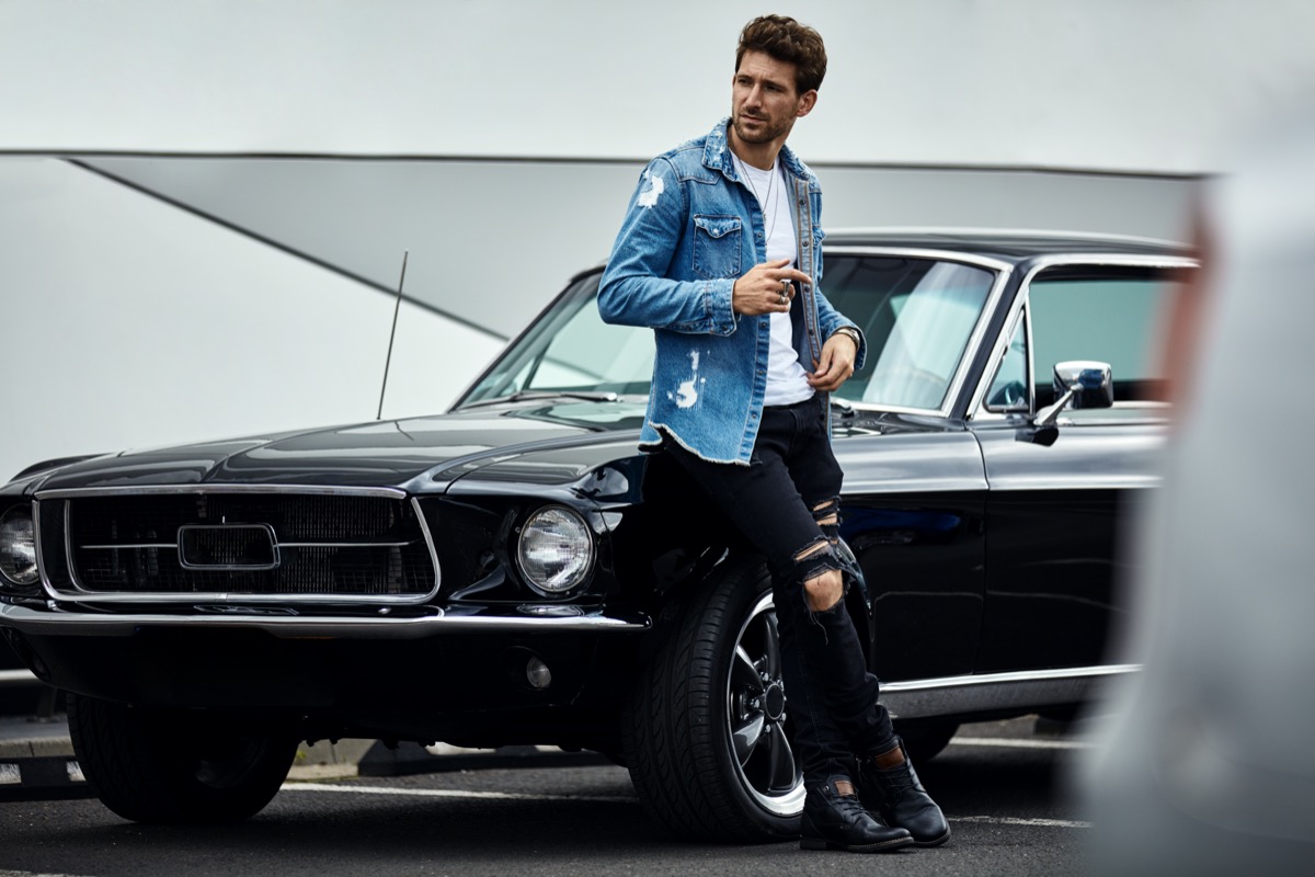 middle aged man wearing jeans and denim jacket and leaning on muscle car