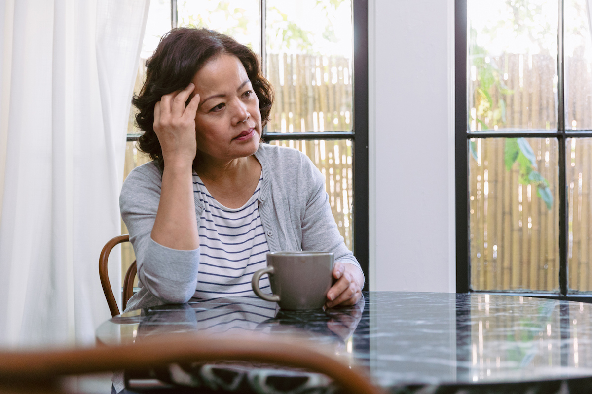 Upset senior woman drags her hand through her hair while staring out the window in her home. She is sitting at the kitchen table. A coffee cup is in front of her.