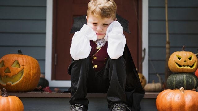 sad kid sitting on stoop, dressed up for halloween with cape, surrounded by pumpkins