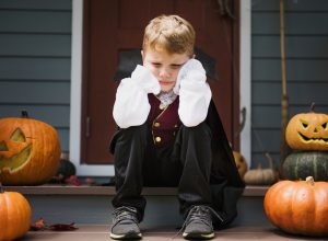 sad kid sitting on stoop, dressed up for halloween with cape, surrounded by pumpkins