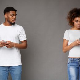 Sad man looking at his girlfriend, who is texting on phone, on gray background