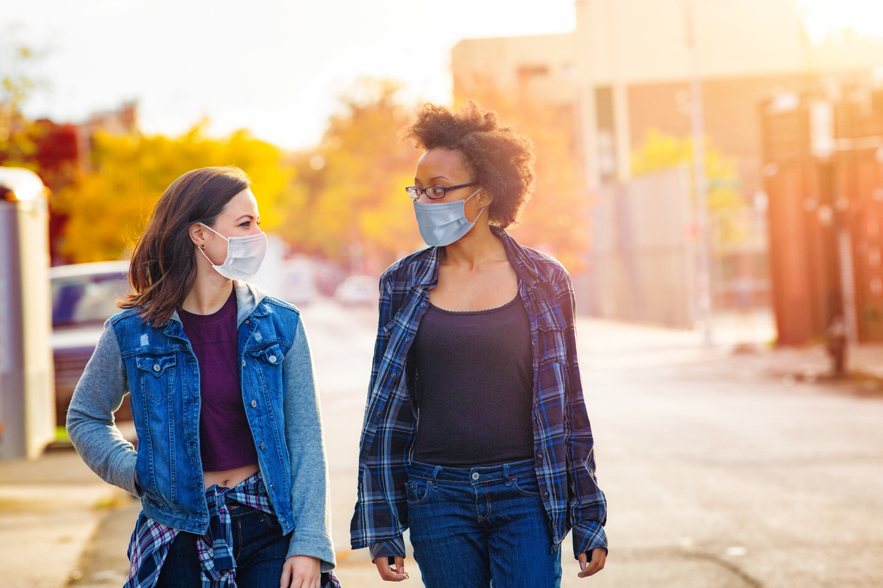 Two young female friends walk in an alleyway while talking to each other and wearing face masks.