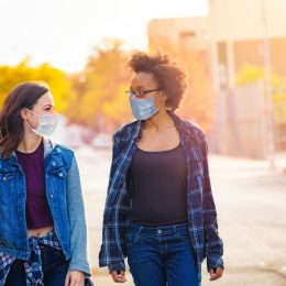 Two young female friends walk in an alleyway while talking to each other and wearing face masks.