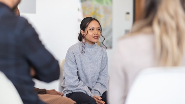 A group of adults are attending a group therapy session. The attendees are seated in chairs arranged in a circle. A mixed-race millennial woman is talking. The other attendees are listening to her speak.