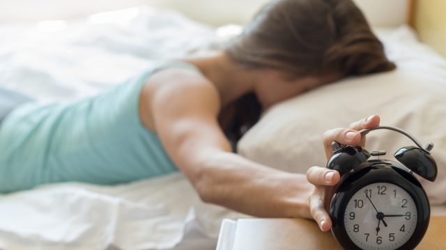 A tired woman in bed hits her alarm clock to stop it from going off.
