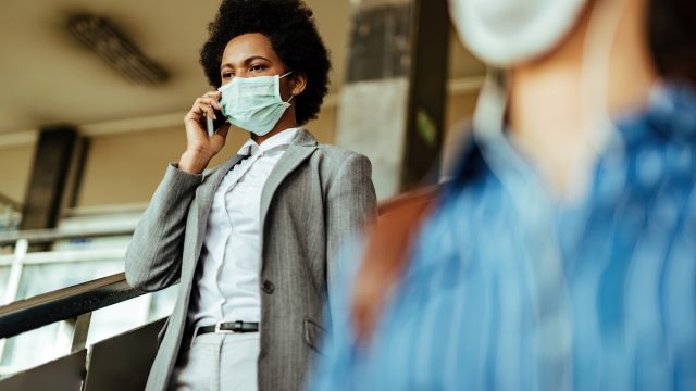 Black woman dressed professionally walks through airport while wearing a mask and talking on the phone