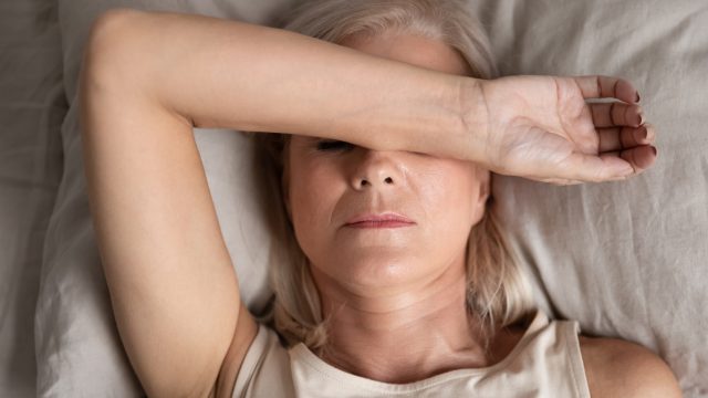 An older woman lying in bed covering her eyes with her arm because she is fatigued.