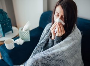 A young woman sitting on the couch wrapped in a blanket and blowing her nose, suffering from flu symptoms.