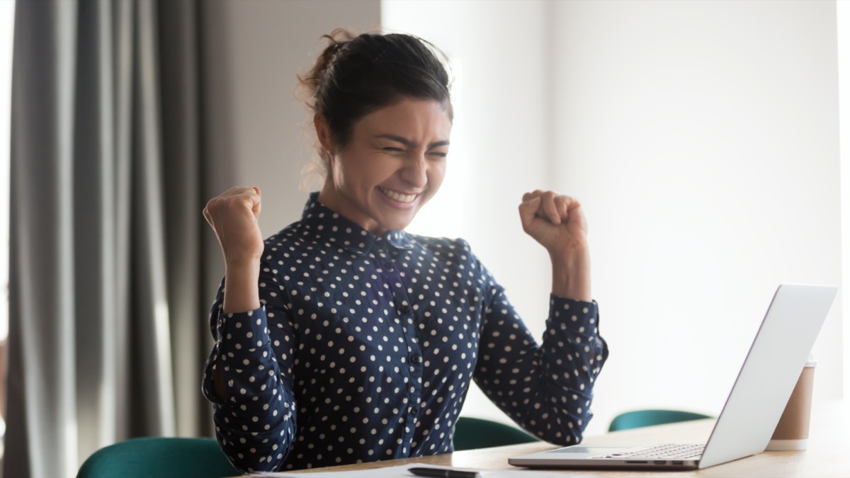 young woman sitting at table looking at laptop, looking exciting and holding her hands up in triumph