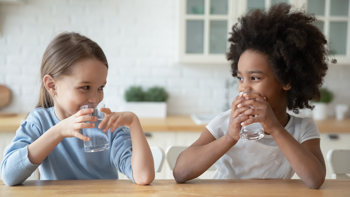 Two young girls sit at a table drinking glasses of water while smiling at each other.