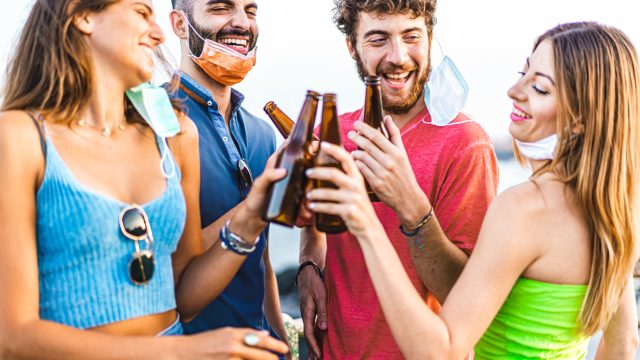 A group of four young men and women cheers beer bottles together with their face masks hanging off, making it easier to spread coronavirus