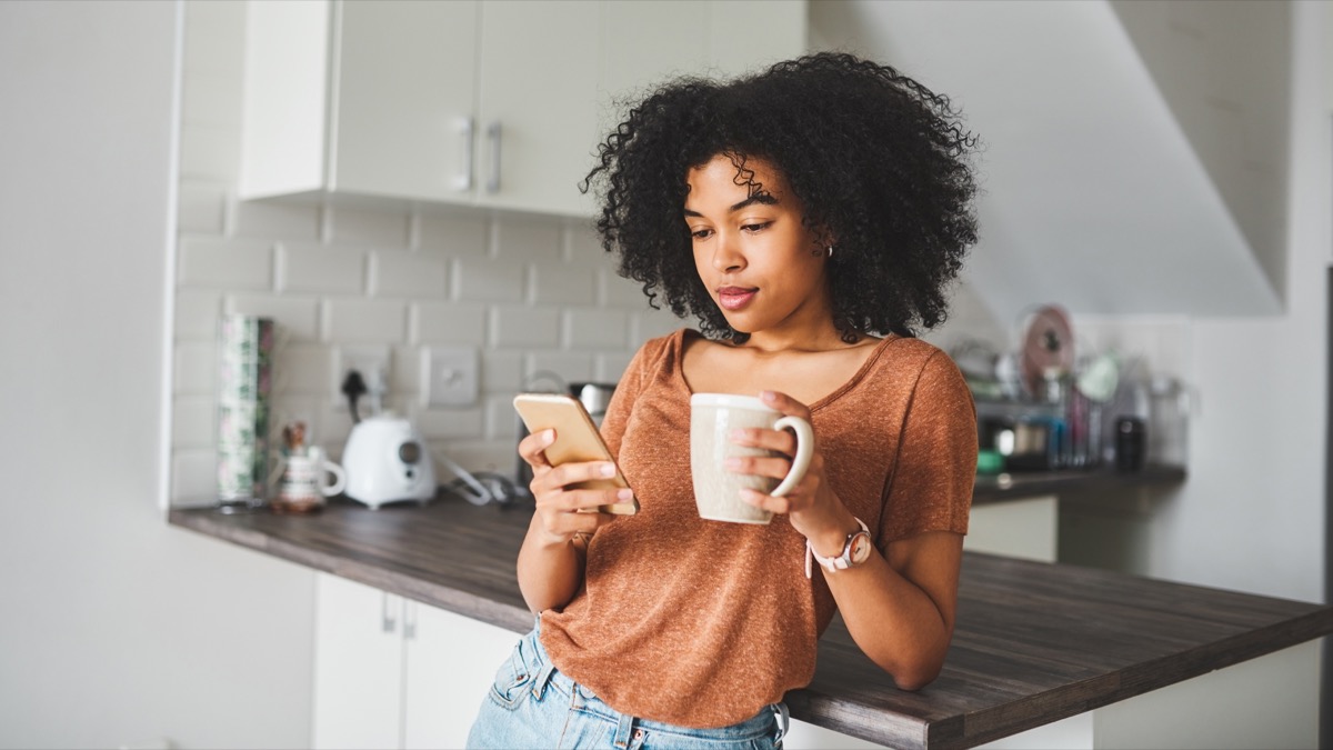 Shot of a young woman using a smartphone and having coffee in the kitchen at home