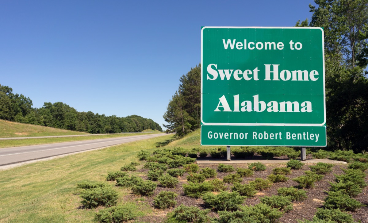 a green "Welcome to Sweet Home Alabama" sign in front of green plants and off a highway