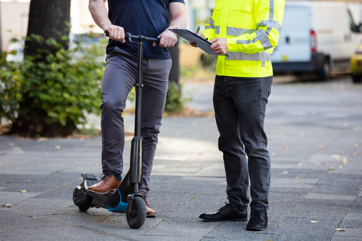 Officer Holding Clipboard In Hand Standing With Man Riding Electric Scooter On City Street