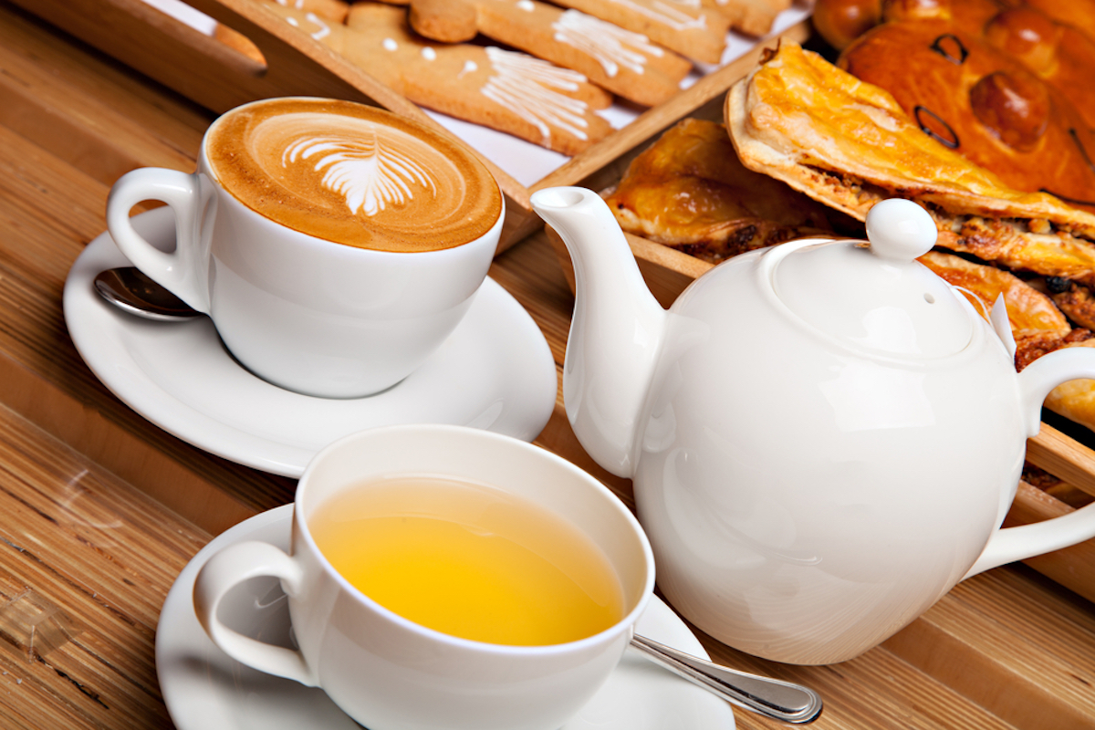 closeup of wooden table with two saucers, one of coffee and one of green tea, with kettle and pastries behind it