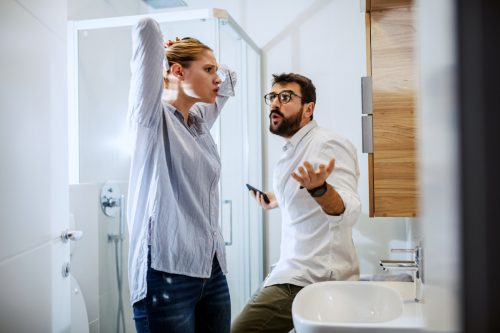 Eine Frau, die sich vor dem Spiegel die Haare frisiert, während der Mann im Badezimmer auf dem Waschtisch sitzt, genervt von ihr guckt und gestikuliert