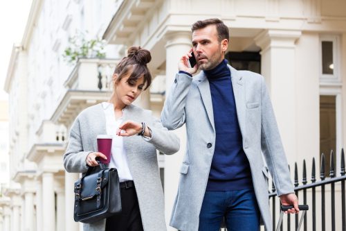 Cuplu ocupat mergând pe strada orașului, femeie ținând cafea și uitându-se la ceas în timp ce bărbatul vorbește la telefonul mobil