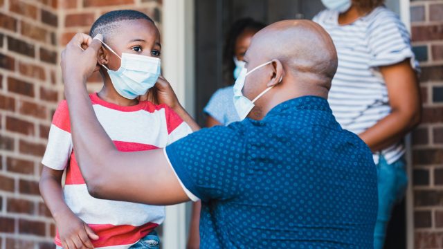 A young Black father puts a face mask on his young son as they leave their house