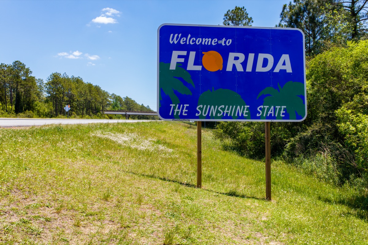 a blue "Welcome to Florida" sign within green grass and in front of trees off of the highway