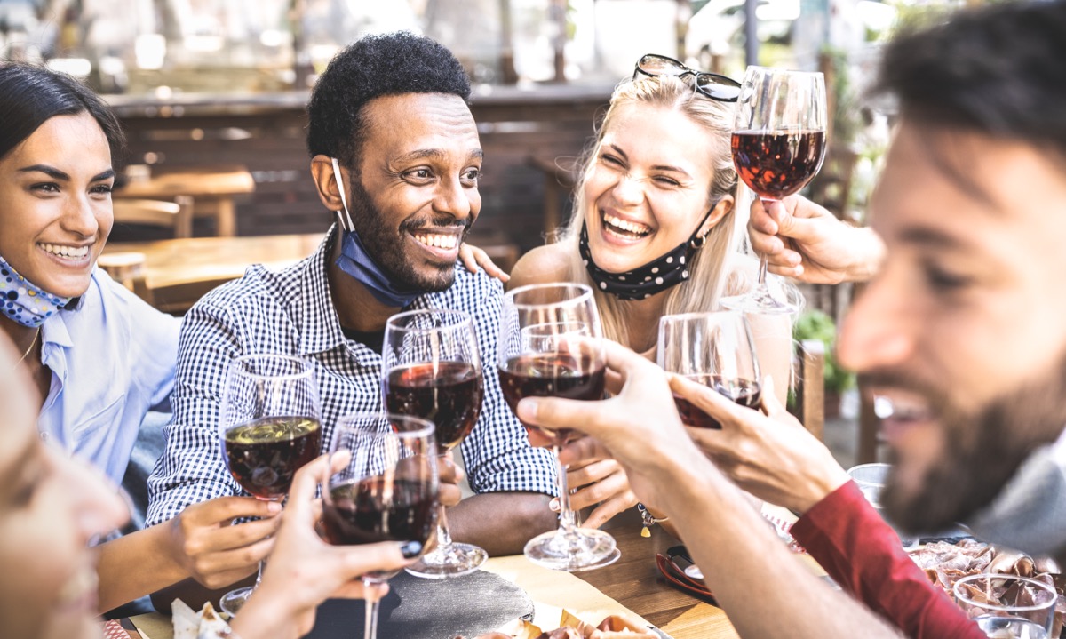Friends toasting red wine at outdoor restaurant bar with open face mask - New normal lifestyle concept with happy people having fun together on warm filte