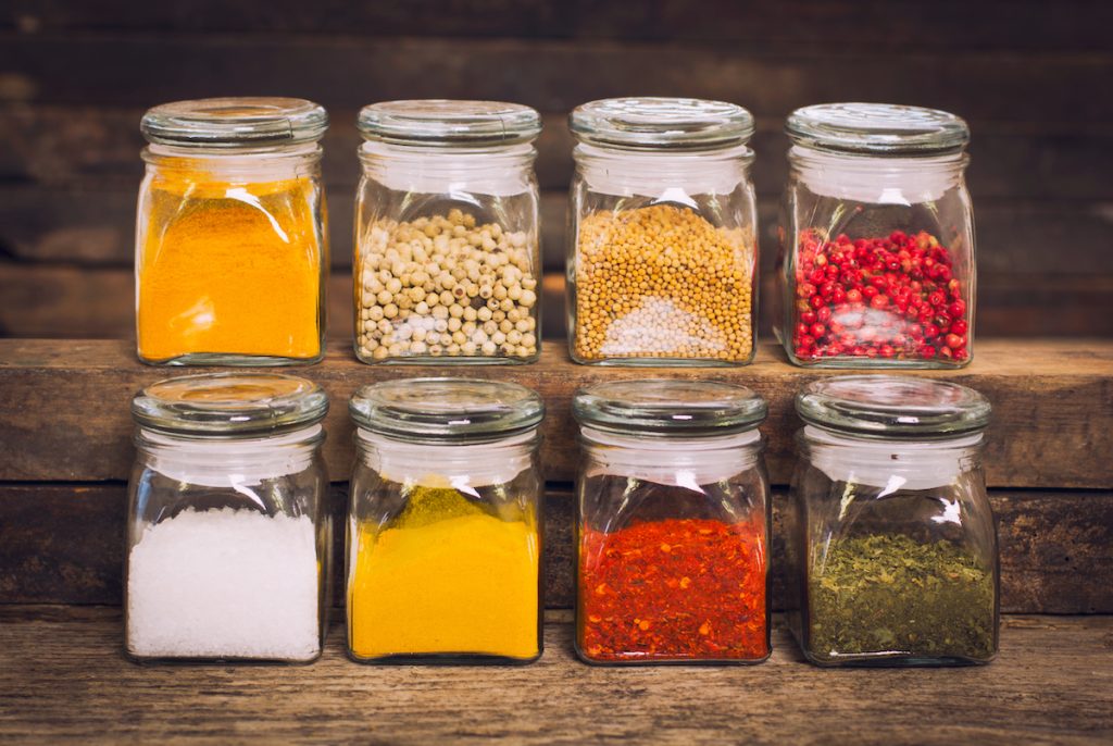 eight small glass jars of spices, in yellow, red, green, and white colors, on natural wood shelves