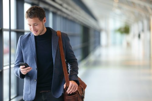 Omul de pe smartphone - tânărul om de afaceri de la aeroport.  Un om de afaceri urban profesionist care folosește un smartphone zâmbind fericit în interiorul clădirii de birouri sau al aeroportului.  Bărbat frumos purtând o jachetă de costum în interior.