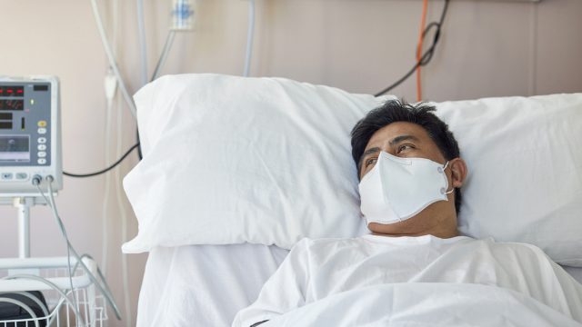 Portrait of male patient in early 40s looking away from camera while lying in hospital bed wearing protective face mask and recovering from coronavirus.