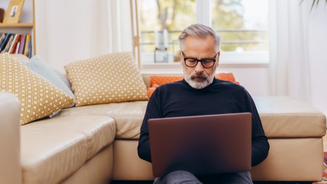 Senior man sitting using a laptop at home relaxing on the floor of the living room leaning against the sofa