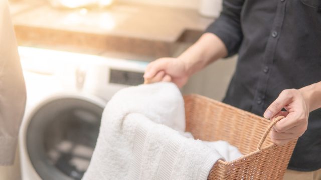 Male hand holding wooden laundry basket with white towel inside near washing machine in laundry room. Home living concept