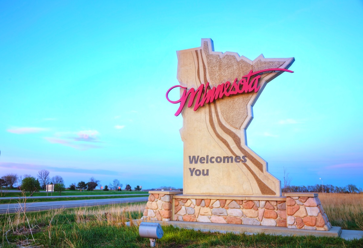 a beige "Minnesota Welcomes You" sign within grass and off the road