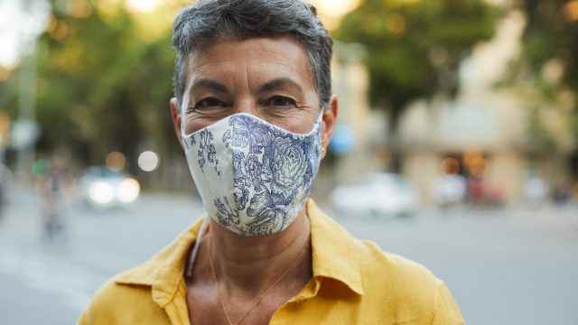 A middle-aged woman in a yellow shirt smiles while wearing a cloth face mask