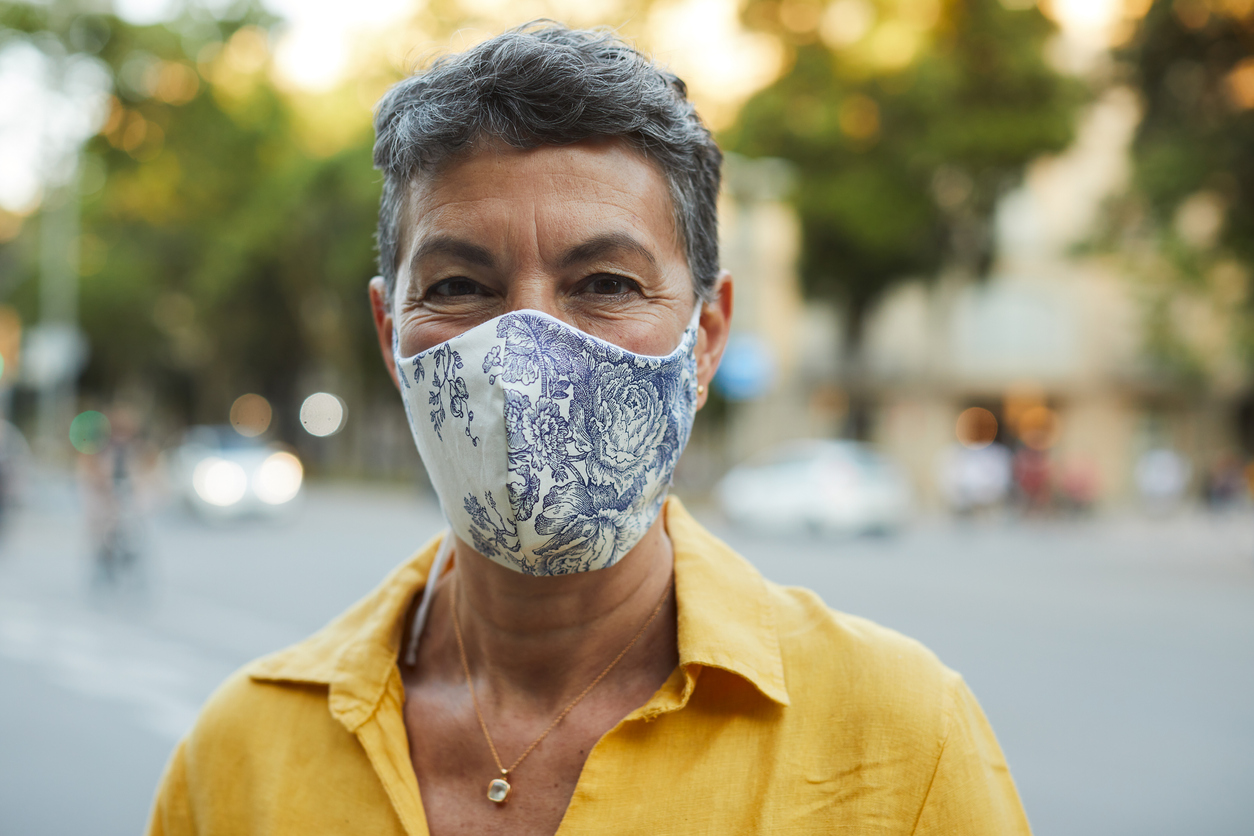 A middle-aged woman in a yellow shirt smiles while wearing a cloth face mask