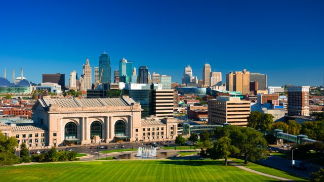 The skyline of Kansas City Missouri with a deep blue sky in the background and Union Station, a fountain, and Penn Valley Park in the foreground.