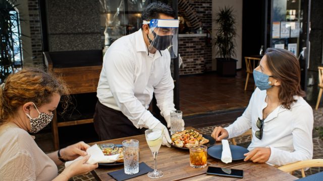 A waiter at a high-end restaurant wearing a face mask and face shield approaches the table carrying a tray, serving to women who are seated and wearing face masks.
