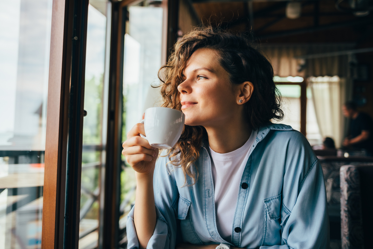 A smiling young woman drinking coffee while looking out the window.