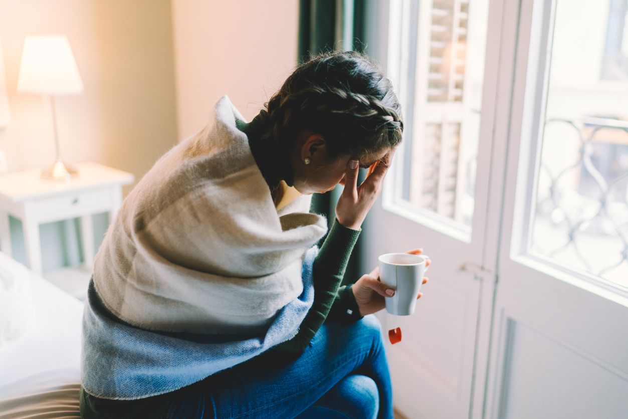 A young woman sitting on her bed wrapped in a blanket while holding a mug of tea feeling symptoms of flu or coronavirus
