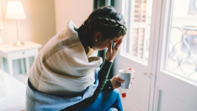A young woman sitting on her bed wrapped in a blanket while holding a mug of tea feeling symptoms of flu or coronavirus