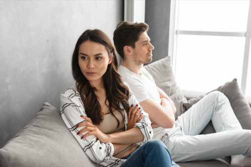 young man and woman sitting on couch arguing with arms crossed