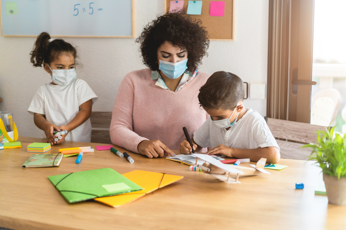 child care provider helping two young children with an art project during coronavirus pandemic; all three are wearing masks