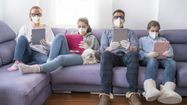 A mother, father, and their two daughters sit on a couch using tablets and devices while wearing face masks.